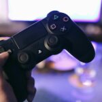 Person holding a gaming controller, blurred console screen in background.