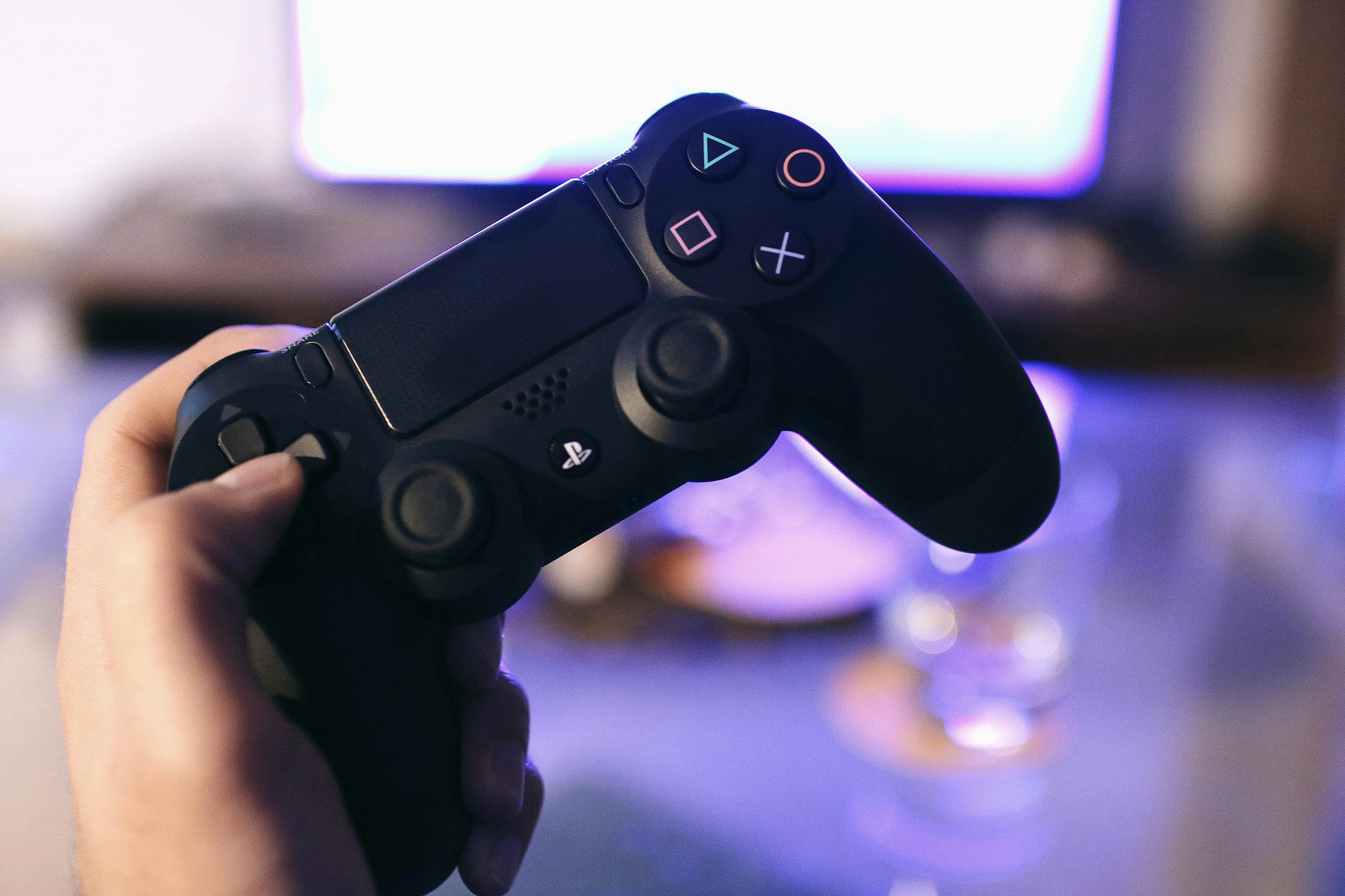 Person holding a gaming controller, blurred console screen in background.